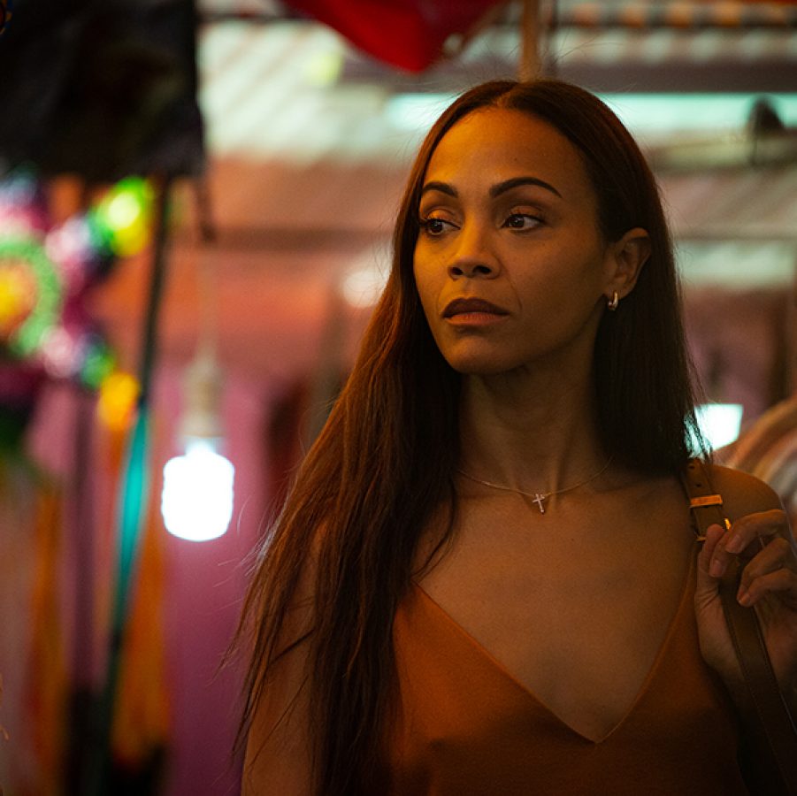 In a scene from the film Emilia Pérez, a woman, Rita (Zoe Saldaña), looks tense as she walks through an illuminated market street at night. 